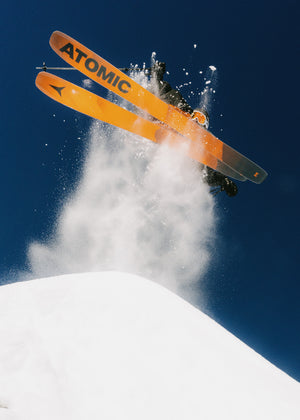 Skier carving through fresh powder with Atomic skis, creating a spray of snow against a clear blue sky, Teton Village, Wyoming – Nomad Sports.