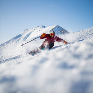 Skier in red jacket making a sharp turn on a snowy mountain slope, spraying powder under a clear blue sky on Kastle skis in Teton Village, Wyoming – Nomad Sports.