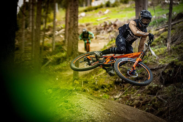 Mountain biker in full gear riding a Pivot Firebird bike on a forest trail, taking a jump with another rider in the background, Teton Village, Wyoming – Nomad Sports.