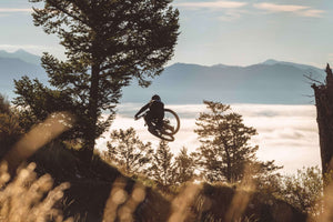 Mountain biker catching air on a scenic trail in Teton Village, Wyoming, with a misty forest and mountains in the background at sunrise – Nomad Sports.